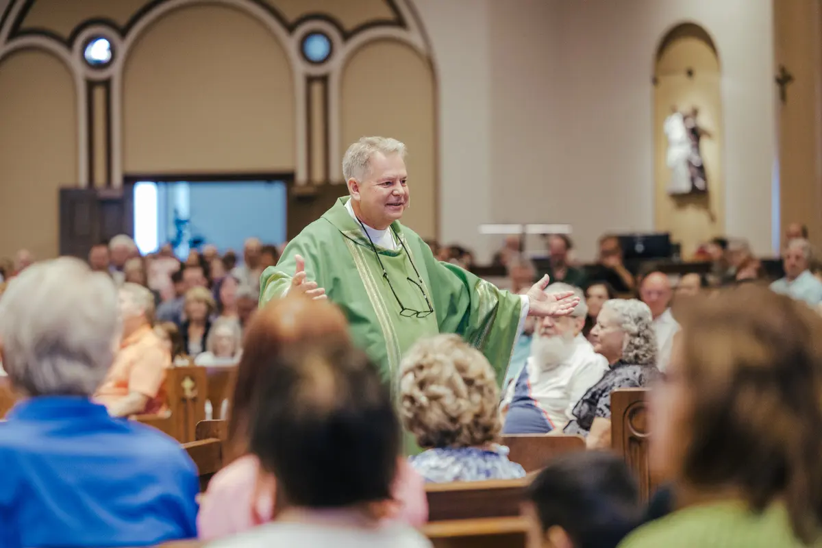 A priest in green vestments extends his arms among the parish congregation.